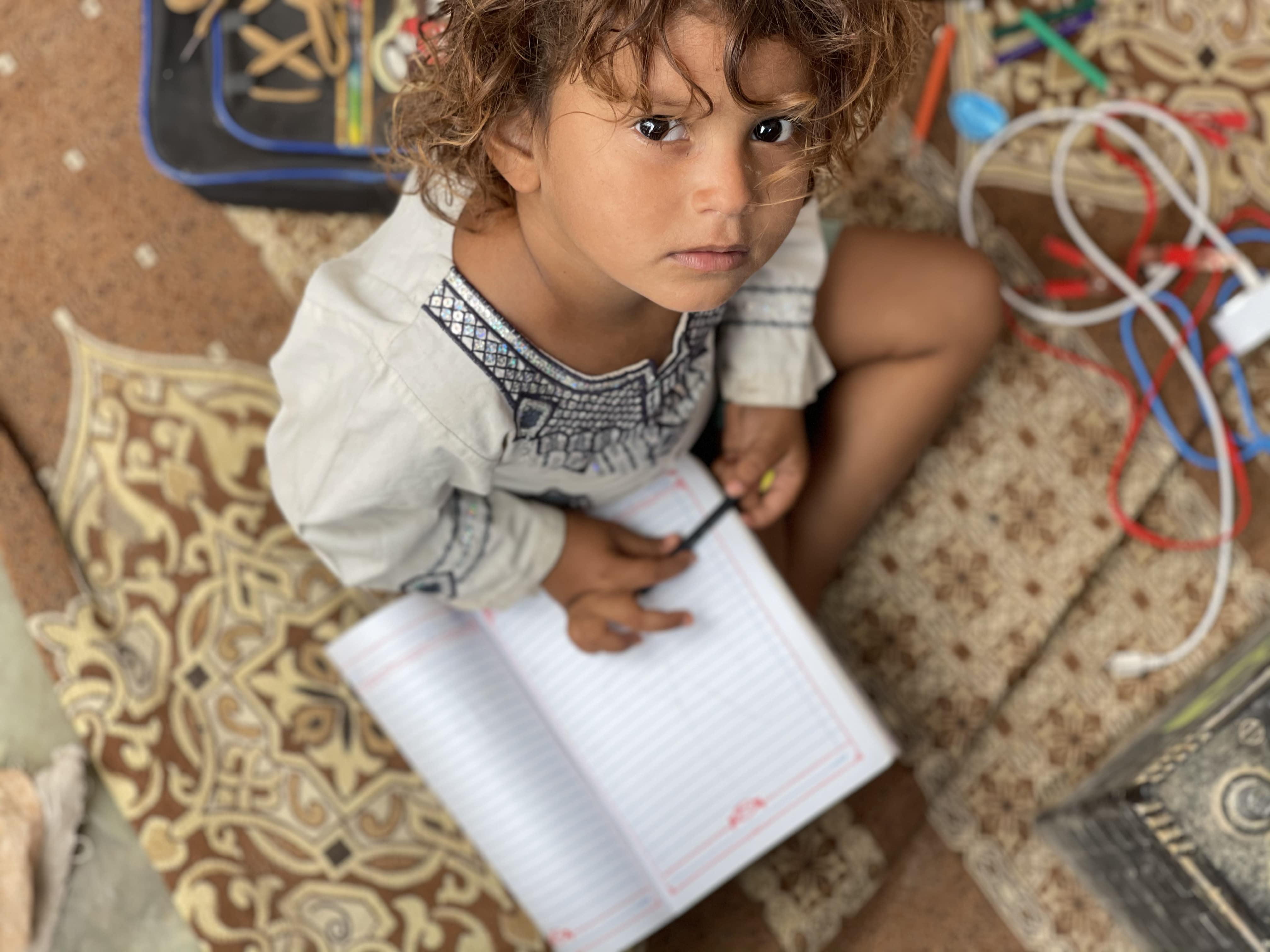 Girl with notepad looks at camera. Displaced Yemeni girl, Umaimah, practices writing in her makeshift shelter.