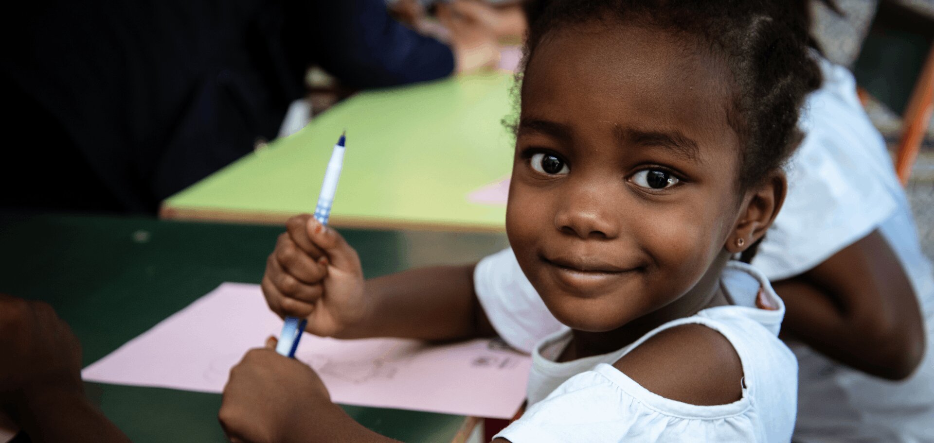 An eight-year-old Tunisian girl sits at a school desk holding a pen and smiling at the camera