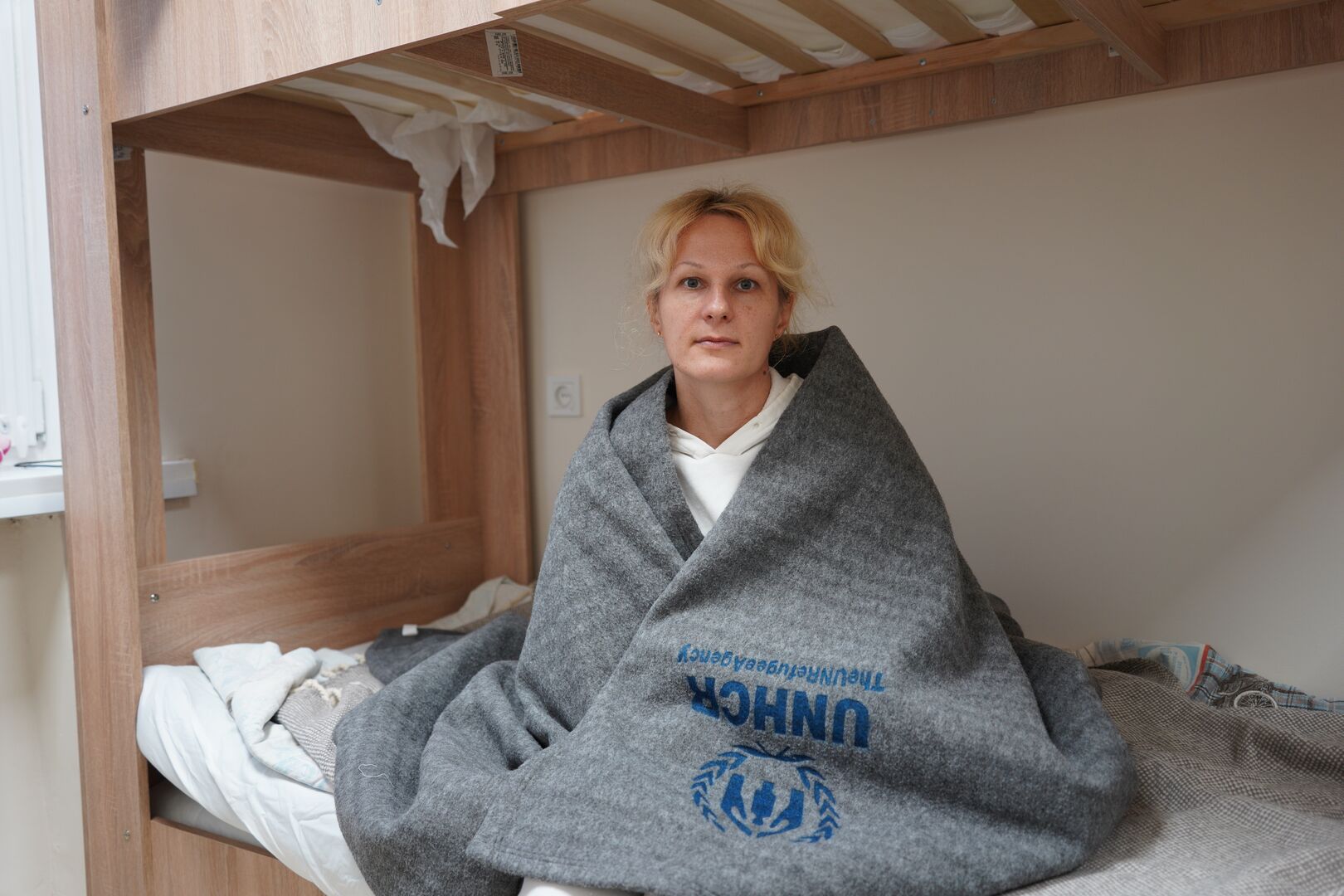 A blond-haired woman sits on a wooden bed wrapped in a grey UNHCR blanket