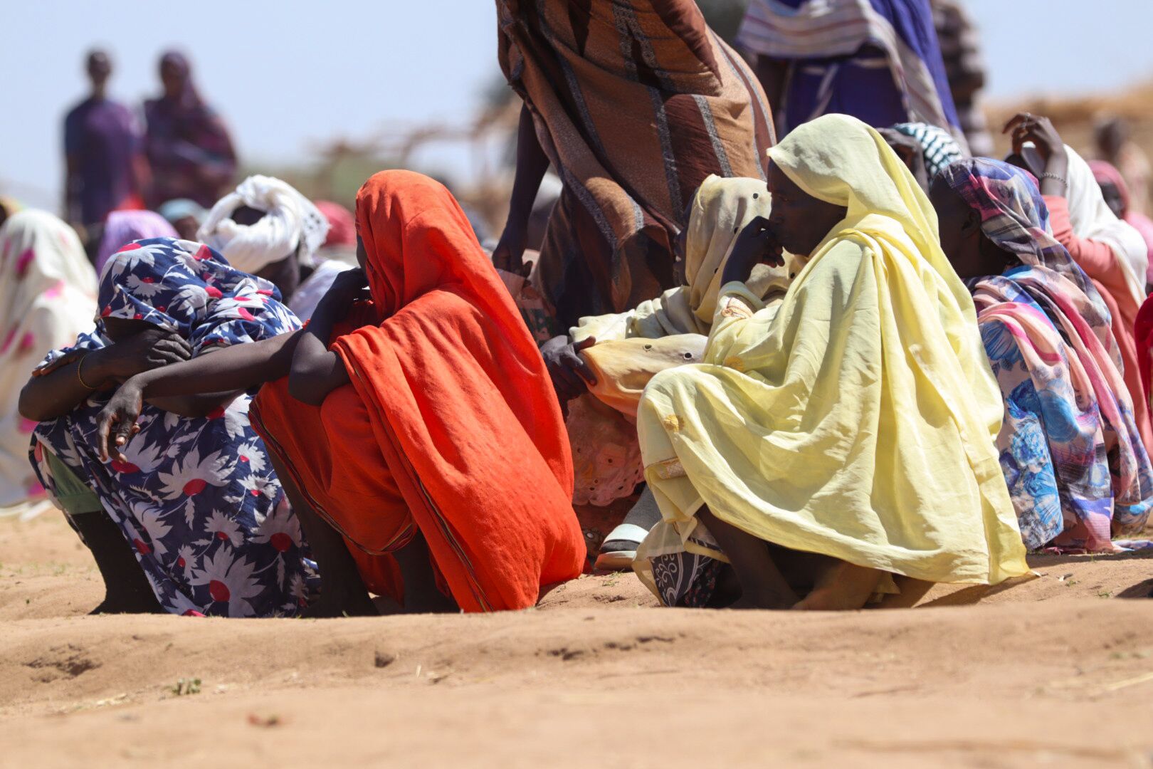 A group of Sudanese women sit on bare ground. They wear brightly coloured shawls, some with their heads resting in their arms.